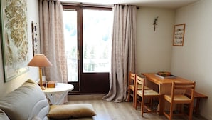 A cozy living room with a sofa, a dining table with chairs, a lamp, and a window with curtains.