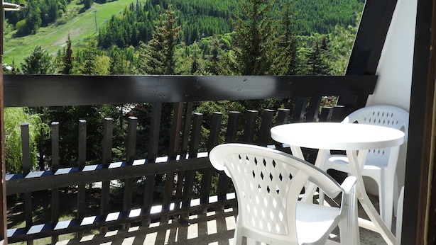 A balcony with a view of a forested valley and mountains. There are two white plastic chairs and a small round table.