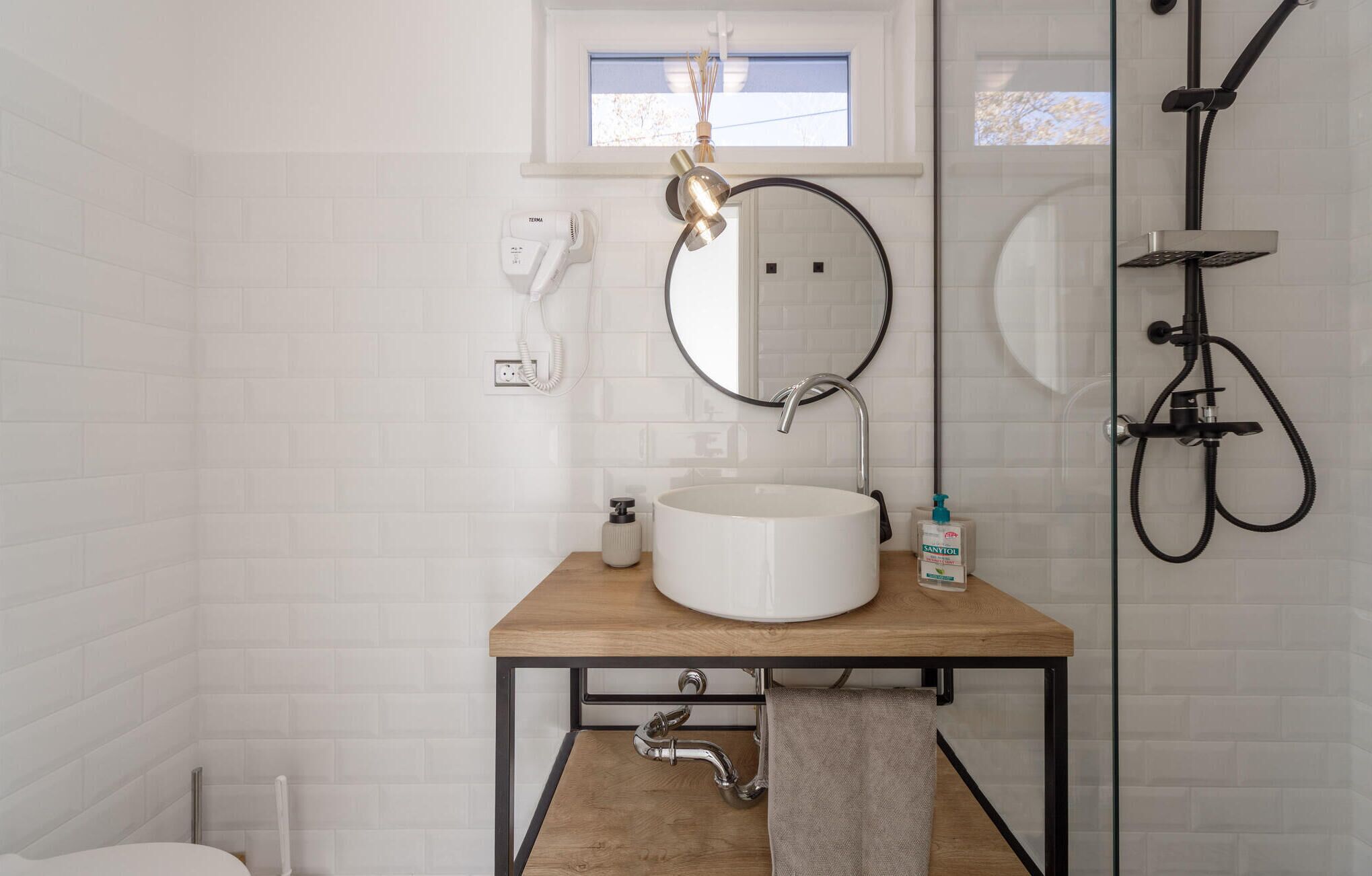 Un baño con un lavamanos blanco, un espejo redondo, un secador de pelo de mano y una ducha con mampara de vidrio.