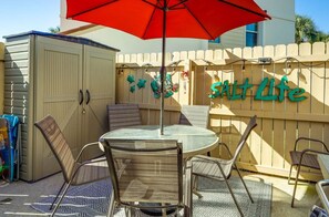 Een terras buiten met een ronde tafel en vier stoelen, een rode parasol en een hek met decoratieve details.