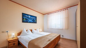 Quarto com cama de madeira, roupa de cama branca, mesa de cabeceira com abajur, pintura na parede e janela com cortinas leves.