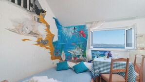 Ett rum med en väggmålning som föreställer marint liv, en säng med blått sänglinne, ett bord med en rutig duk och ett fönster med utsikt över havet.