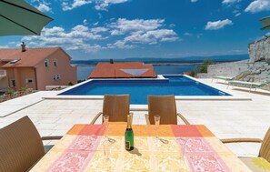 A poolside area with a table set for two, two wicker chairs, and a bottle of champagne. The pool is rectangular with a tiled deck surrounding it. - Amazing Home in Dramalj With Wifi, 2 Bedrooms and Outdoor Swimming Pool (Crikvenica)