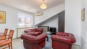 A modern living room with a sectional sofa, a dining area with wooden chairs, a television, and a large window with blinds. - Stunning Apartment in Pula With Wifi and 2 Bedrooms (Pula)