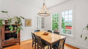 A dining room with a wooden table and chairs, a chandelier, a cabinet with plants, and a window with a view of greenery. - Mountain View Cottage | Wine, Hiking & Dog Friendly (Afton)