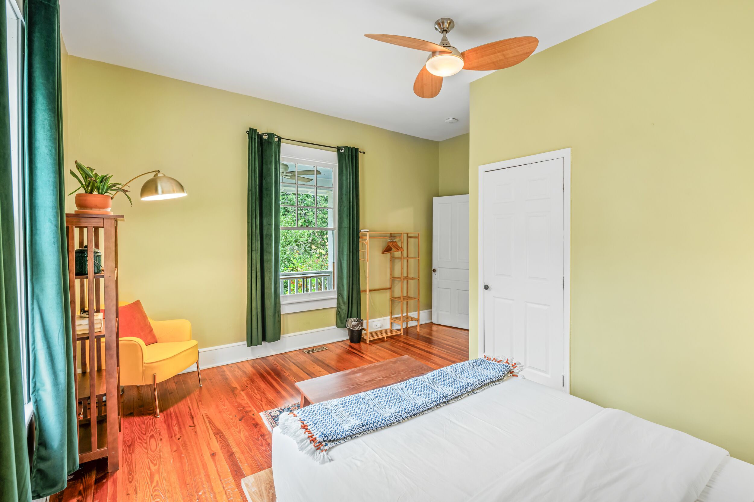 Une chambre à coucher avec un lit, un ventilateur de plafond, une fenêtre avec des rideaux, une chaise et une petite table sur laquelle est posée une plante.
