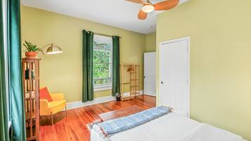 Une chambre à coucher avec un lit, un ventilateur de plafond, une fenêtre avec des rideaux, une chaise et une petite table sur laquelle est posée une plante.