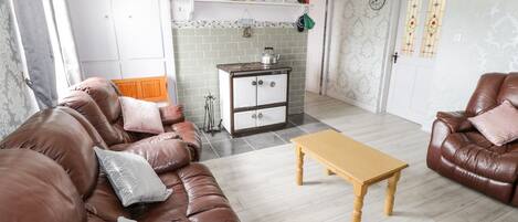 Un salon avec un canapé en cuir marron, une table basse en bois et un coin cuisine équipé d’une cuisinière et de placards.