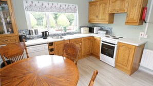 A kitchen with wooden cabinets, a white stove, a sink, and a dining table with chairs. - Sean Bhaile (Glenisland)