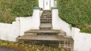 A white house with a front door, a set of concrete steps leading up to the door, and two large, round hedges on either side. - Sean Bhaile (Glenisland)