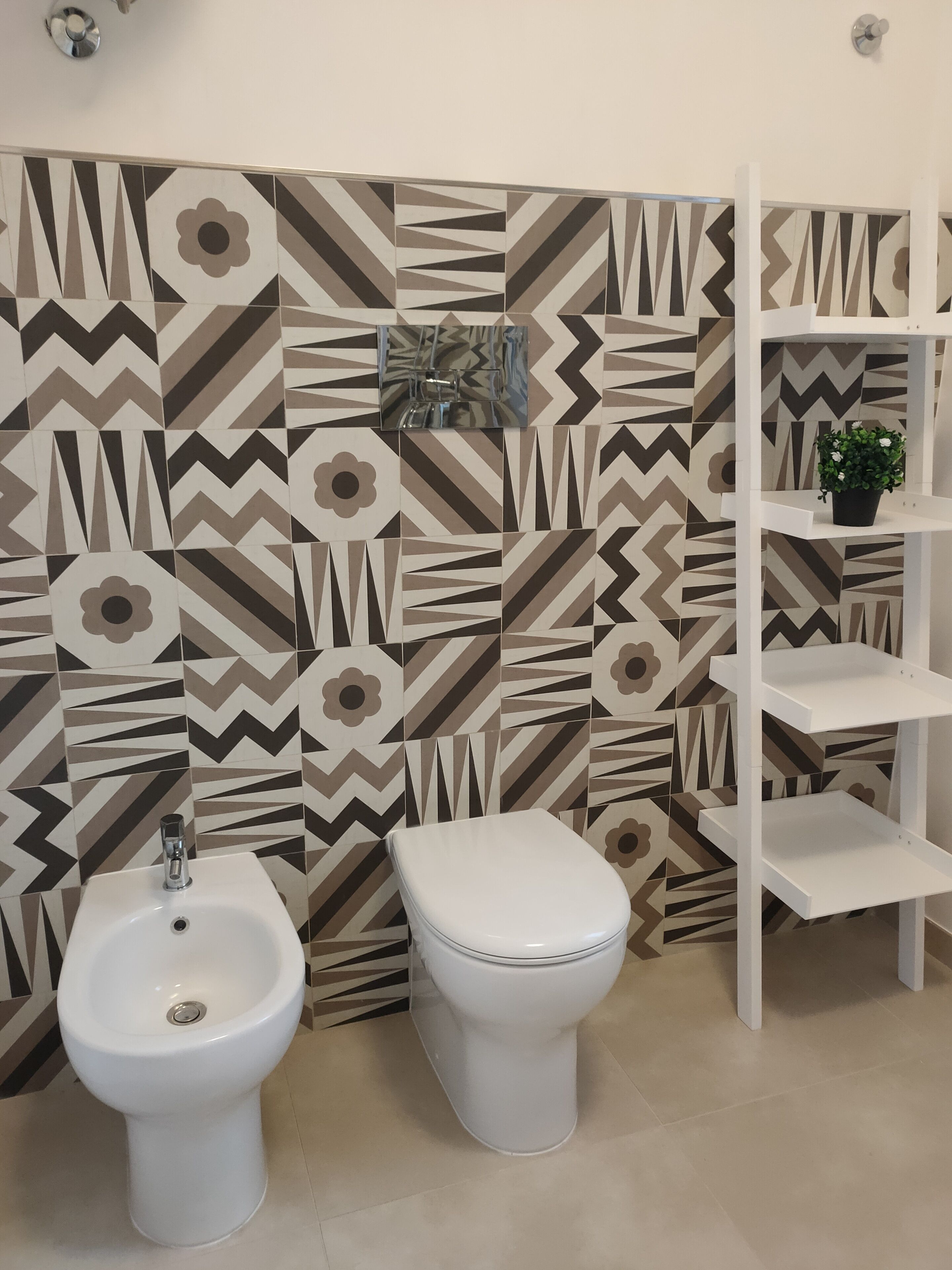Et badeværelse med et flisebeklædt backsplash, en hvid pedestalvask, et toilet og en hvid reol med en potteplante.
