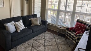 Een woonkamer met een donkergrijze bank, gekleurde kussens, een geruit fauteuil, een kleine ronde tafel met een vissenkom en een uitzicht op gebouwen door grote ramen met jaloezieën.
