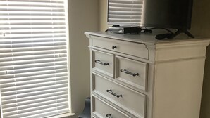Een witte dressoir met laden en een flatscreen televisie erop, geplaatst naast een raam met jaloezieën.