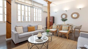 A modern living room with a grey sofa, two round tables, wicker chairs, and a large window with white shutters.