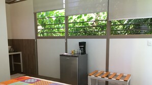 A room with a bed, a small table, a bench, a refrigerator, and a window with a view of greenery. - Eliott Jungle (Jaco)