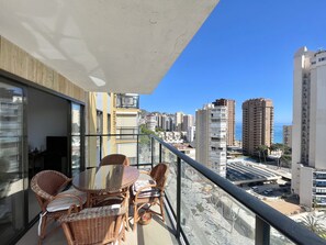 A balcony with wicker furniture and a glass table, overlooking a cityscape with tall buildings and a clear blue sky. - FRONT AL MAR LEVANTE BEACH R079 (Benidorm)