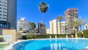 Ein Swimmingpool mit klarem, blauem Wasser, umgeben von einem weißen Beckenrand und einer Hecke. Im Hintergrund sind hohe Gebäude und Palmen zu sehen.