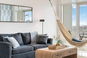 A modern living room with a grey sofa, a wooden side table, a hanging hammock, and a large window with a view.