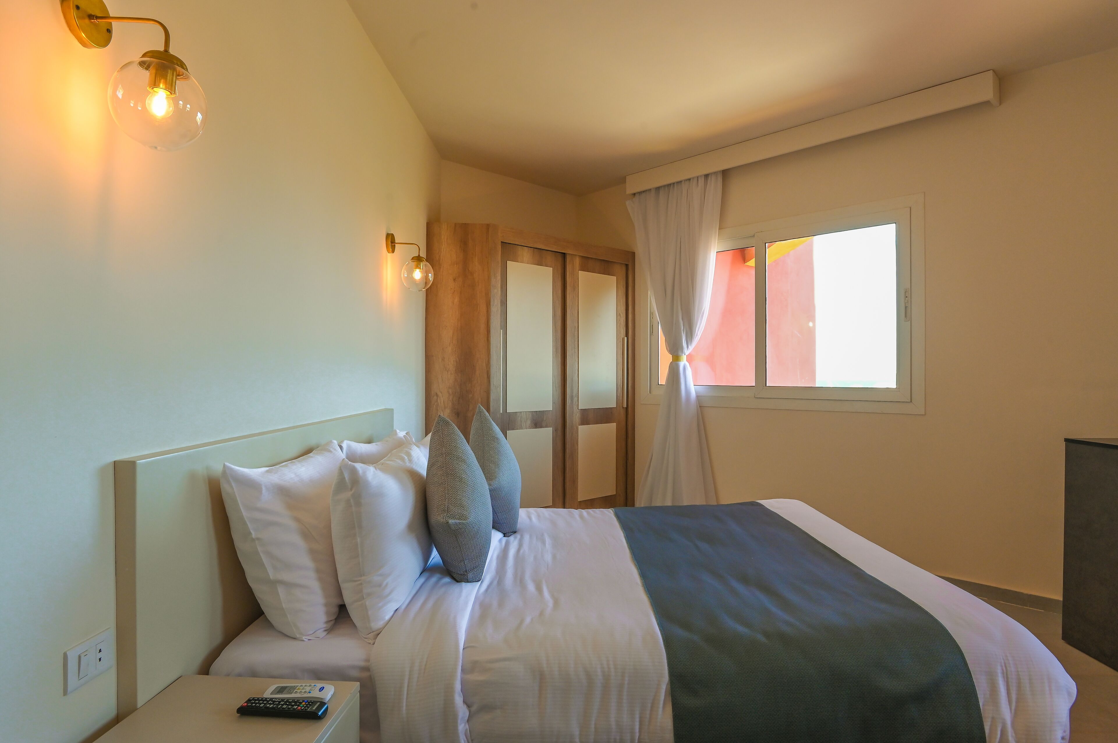 A hotel room with a bed, a nightstand, a remote control, a wall-mounted light fixture, and a window with curtains.