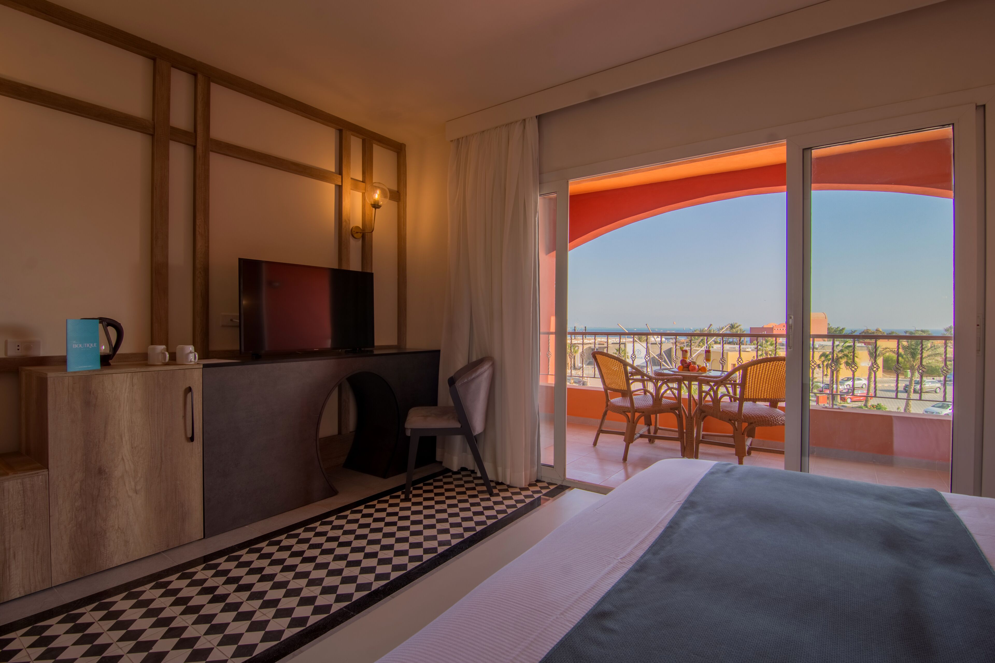 A hotel room with a checkered floor, a bed, a desk with a television, a chair, and a balcony with a view.
