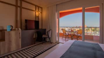 A hotel room with a checkered floor, a bed, a desk with a television, a chair, and a balcony with a view.