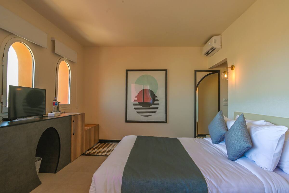 Una habitación de hotel con cama, escritorio con computadora, un cuadro en la pared y ventana de arco ojival.