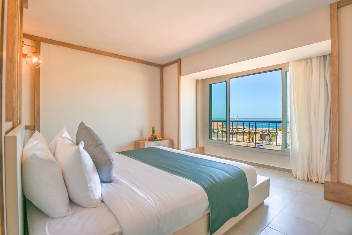 Habitación de hotel con cama, mesita de noche, ventana con vista a la playa y balcón con barandilla.