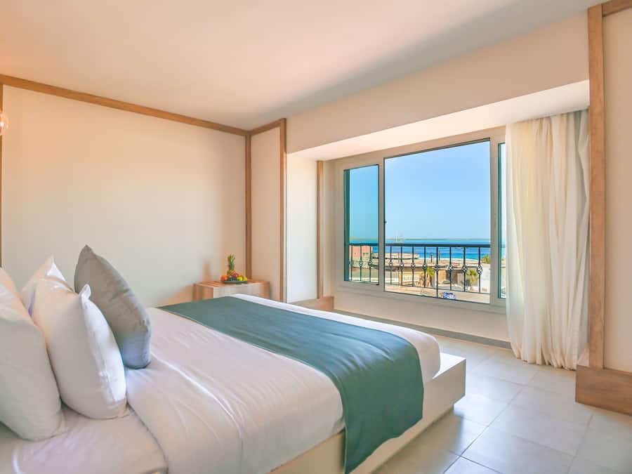 Ein Hotelzimmer mit Bett, Nachttisch, Fenster mit Blick auf den Strand und einem Balkon mit Geländer.