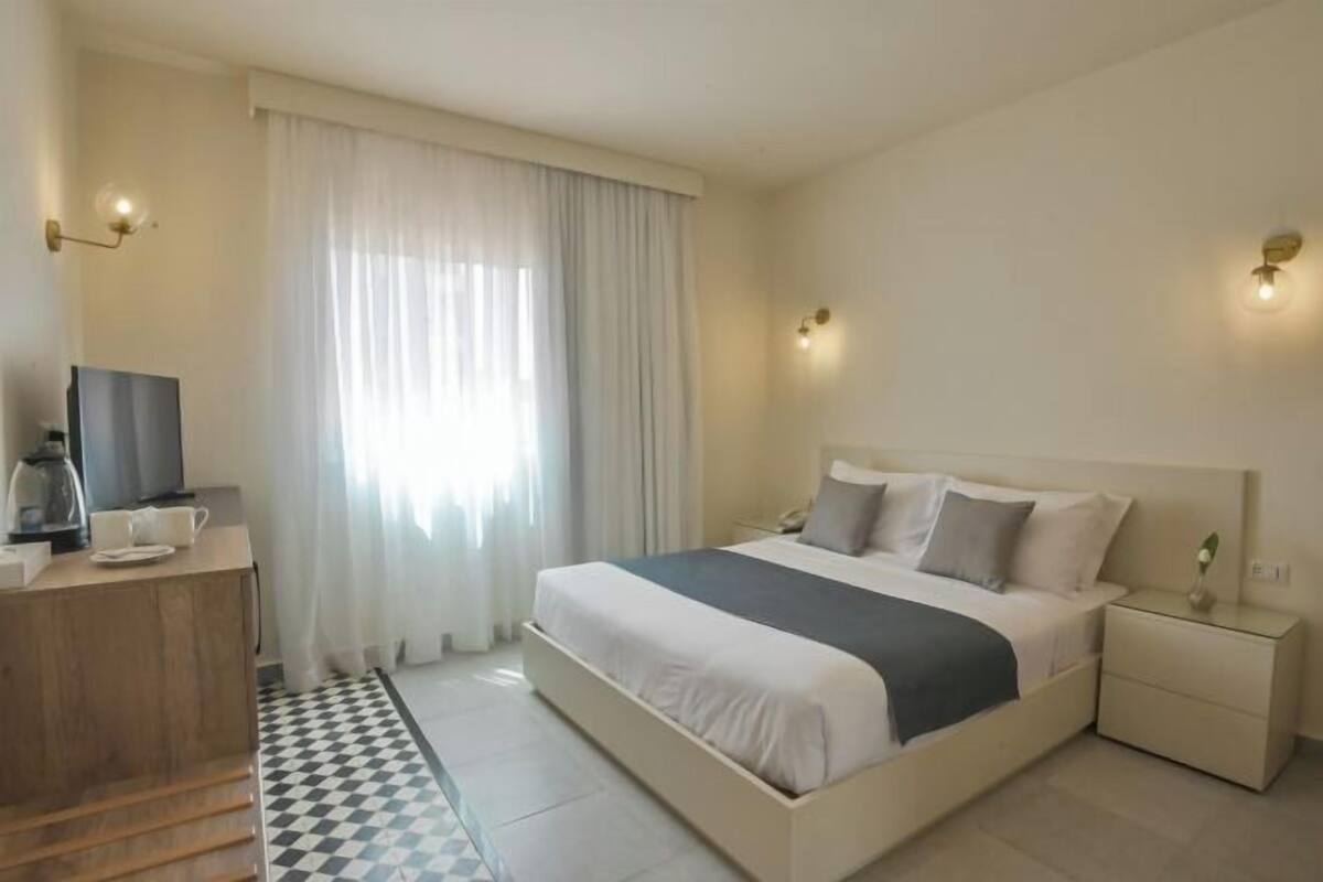 Habitación de hotel con cama, mesitas de noche, escritorio con silla, televisor y ventana con cortinas.