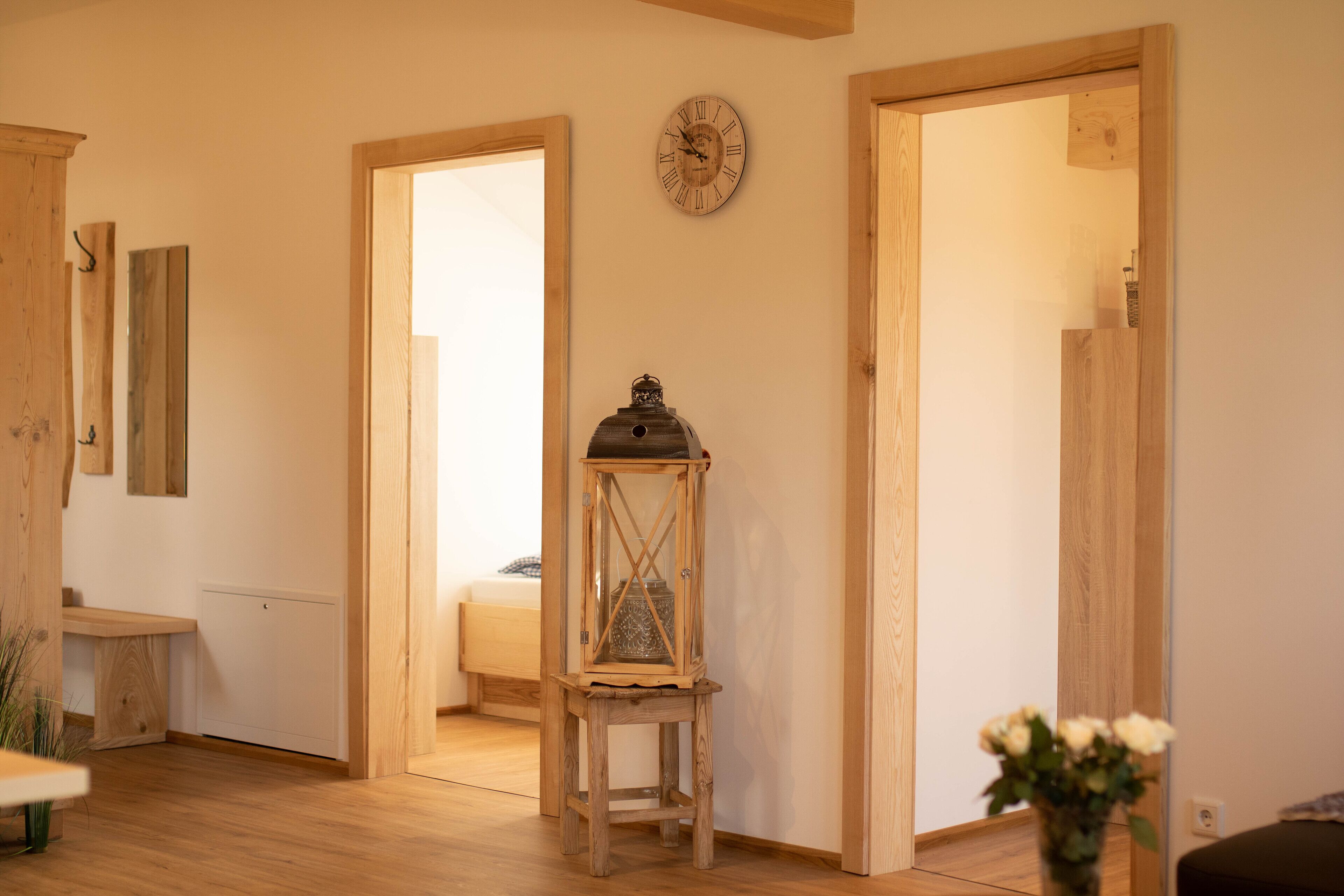 Ein Zimmer mit Holzboden, einem Holztürrahmen, einer Wanduhr und einem kleinen Tisch mit Blumen in einer Vase.