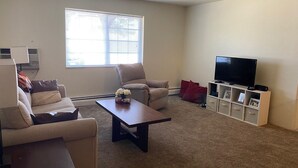 Un salon comprenant un canapé, un fauteuil, une table basse, un meuble pour la télévision et une fenêtre avec des stores.