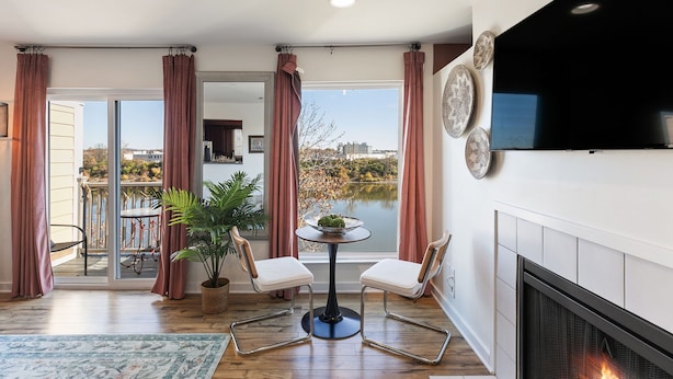 A modern living room with a fireplace, a dining area with two chairs, a small table with a plant, and a view of a lake through large windows. - Downtown Condo with River View (Nashville)