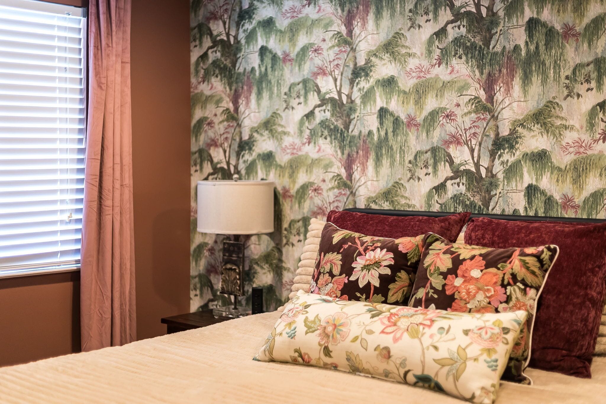 Een slaapkamer met een bed, sierkussens met bloemendessin, een nachtkastje met een lamp en een muur met behang met natuurmotieven.