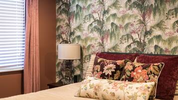 Een slaapkamer met een bed, sierkussens met bloemendessin, een nachtkastje met een lamp en een muur met behang met natuurmotieven.