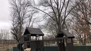 A playground with two wooden playhouses, a slide, and a swing set. - Home on the Green River! (Colona)