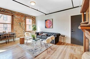 Una sala de estar moderna con pared de ladrillo, un sofá de cuero negro, una mesa de centro de vidrio y un comedor con mesa y sillas de madera.