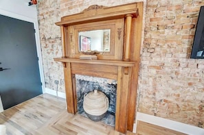 A room with a brick wall, a wooden fireplace with a mirror above it, and a woven basket below. - Vaughn Lofts # 4 (Cincinnati)