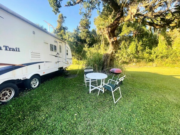A white RV with "On Trail" written on it, parked on a grassy area with a small white table and two chairs.