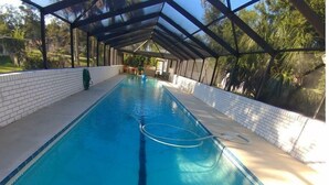 A long, rectangular swimming pool with a clear blue water surface, surrounded by a covered area with a brick wall and a metal roof structure. - RV NSB Sun & Shade Tree, Pool & Picnic RV Vacation (New Smyrna Beach)