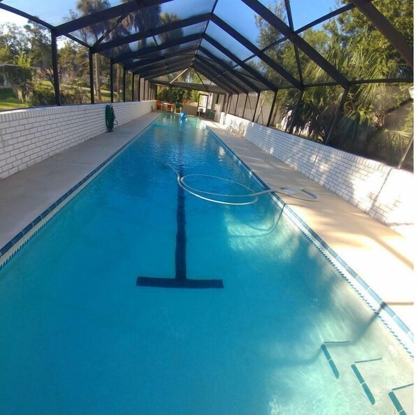 A long, rectangular swimming pool with a clear blue water surface, surrounded by a covered area with a brick wall and a metal roof structure.