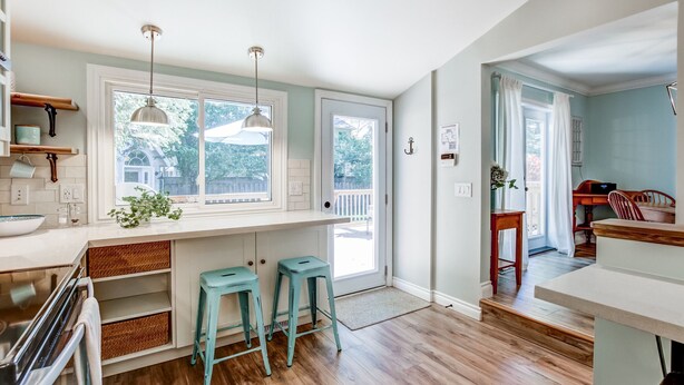 A kitchen with wooden flooring, a white countertop, two bar stools, pendant lights, and a view through large windows. - Blue Pearl, Your Tranquil Oasis (Niagara-on-the-Lake)