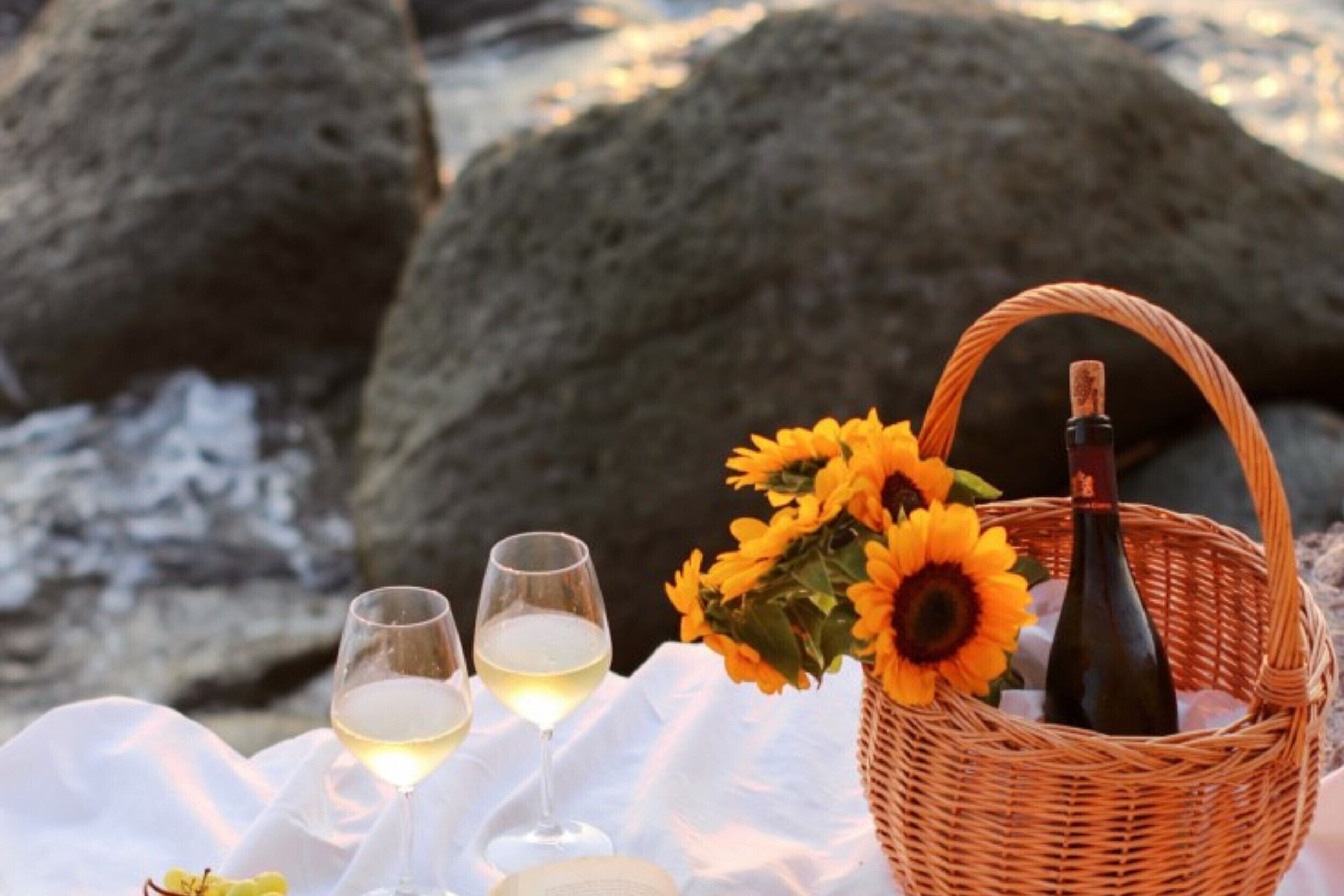 A picnic setup with a wicker basket containing a bottle and sunflowers, two wine glasses, and a bouquet of flowers on a white cloth by rocks.