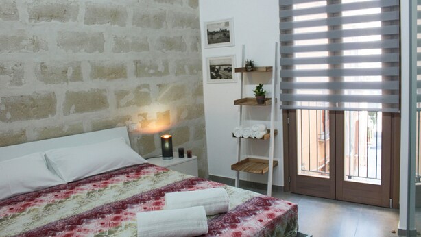 A bedroom with a brick wall, a bed with a floral bedspread, a bedside lamp, a shelf with towels, and a window with blinds. - Suite Torre Pali (Trapani)