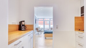 A modern kitchen with a coffee maker, wooden countertop, and white cabinets. A hallway leads to a bedroom with a bed and a desk.