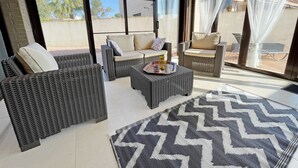 A sunlit patio with wicker furniture, a patterned rug, and a view of buildings through large windows. - CASA DANI (Ampolla)