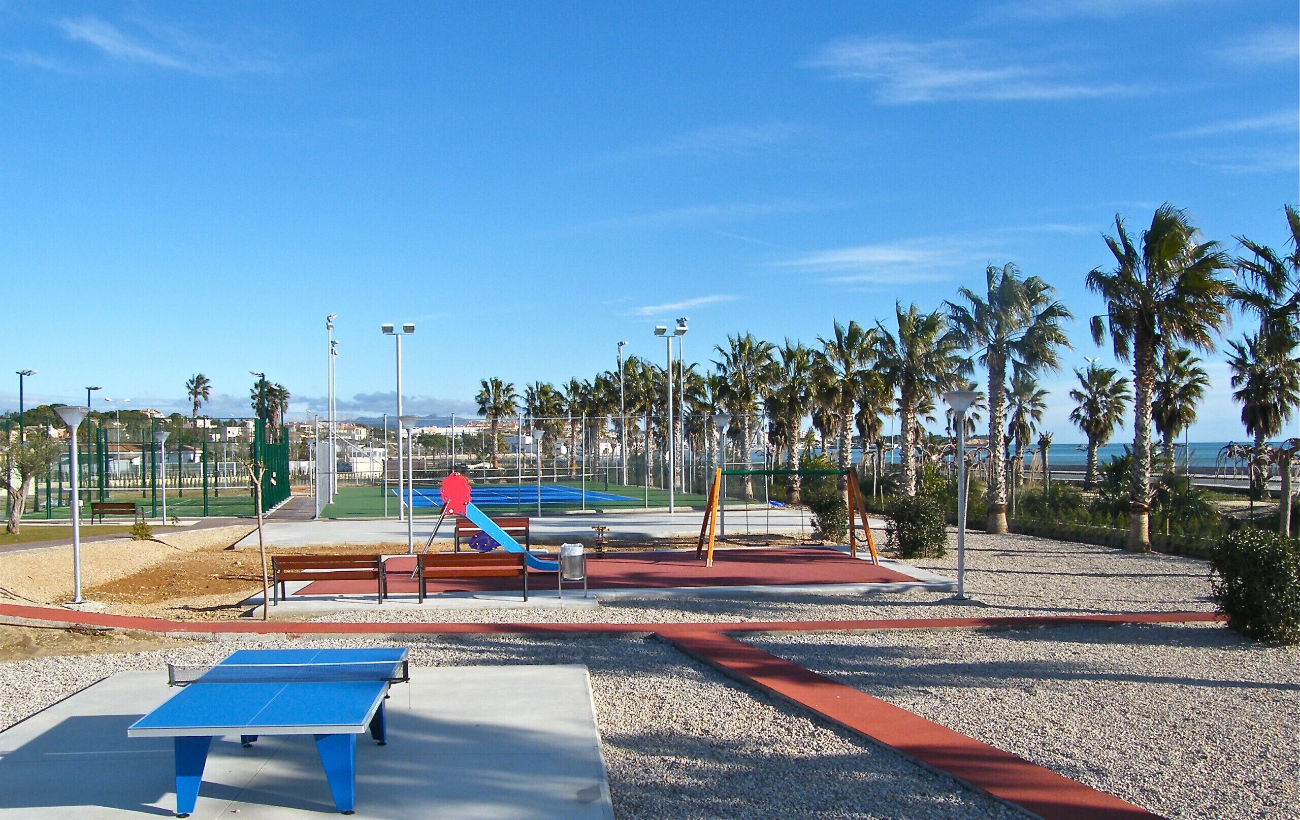 A sports court with a blue table tennis table, a red and blue playground, and palm trees.