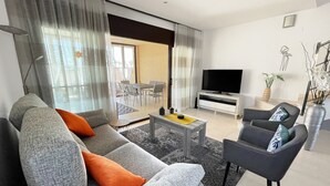 A modern living room with a sofa, armchair, coffee table, TV, and a dining area with chairs and a table. - CASA DANI (Ampolla)