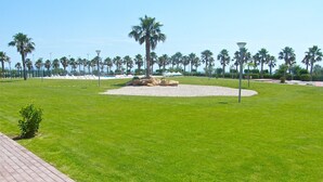 A well-maintained grassy area with palm trees, a circular stone feature, and a paved walkway. - CASA DANI (Ampolla)