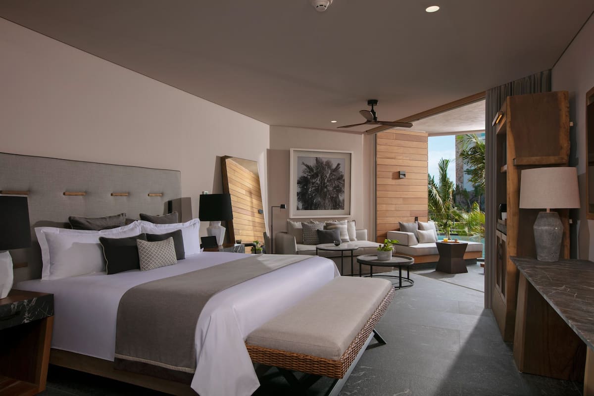 Una habitación de hotel moderna con una cama grande, una zona de estar con sofá y sillones, una mesita y vistas al exterior.