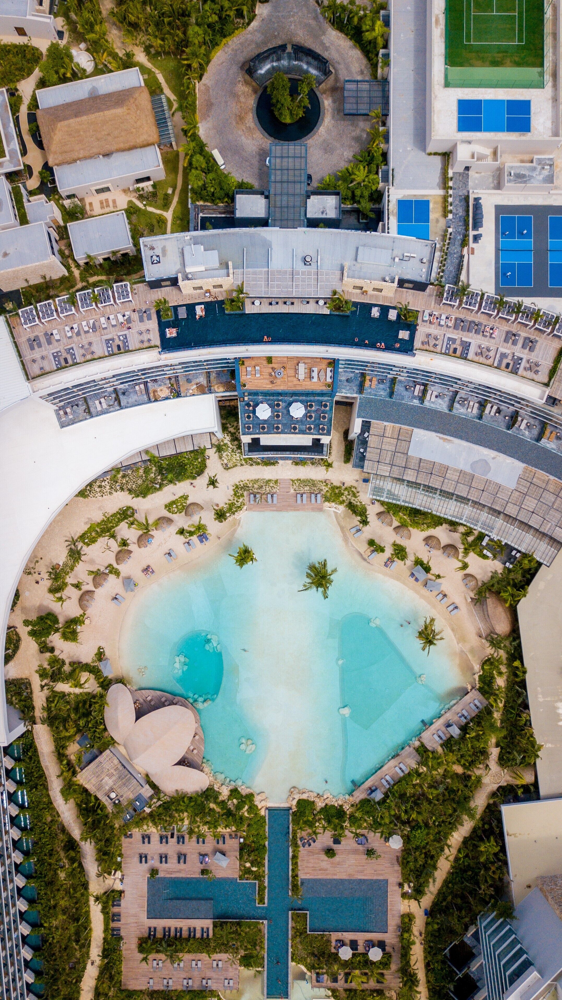 Ovenudblik over et resort med en central pool, flere loungeområder og et cirkulært vandelement.